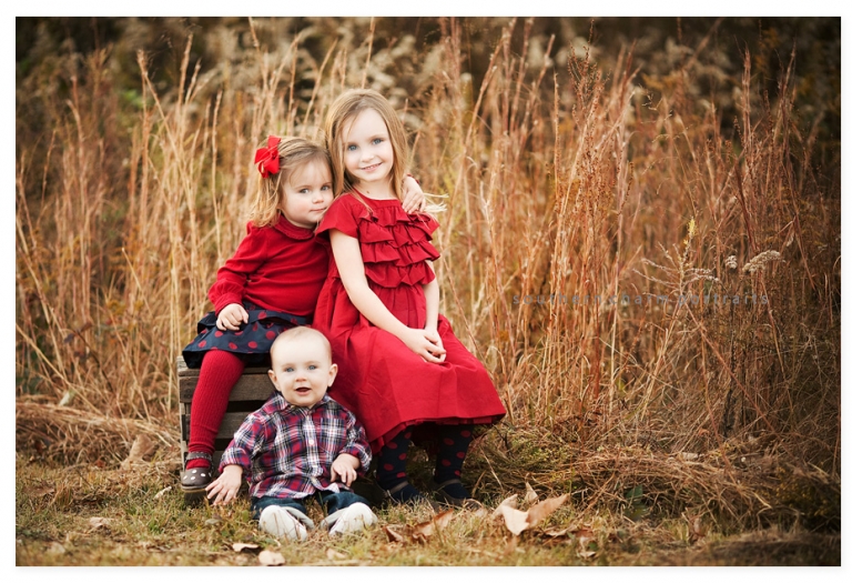 three siblings sitting together on old wooden crate in field of tall grass